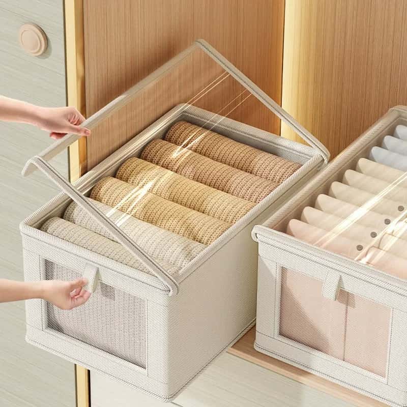 Beige fabric closet storage boxes with clear zippered lids and front windows, holding neatly folded sweaters and shirts.